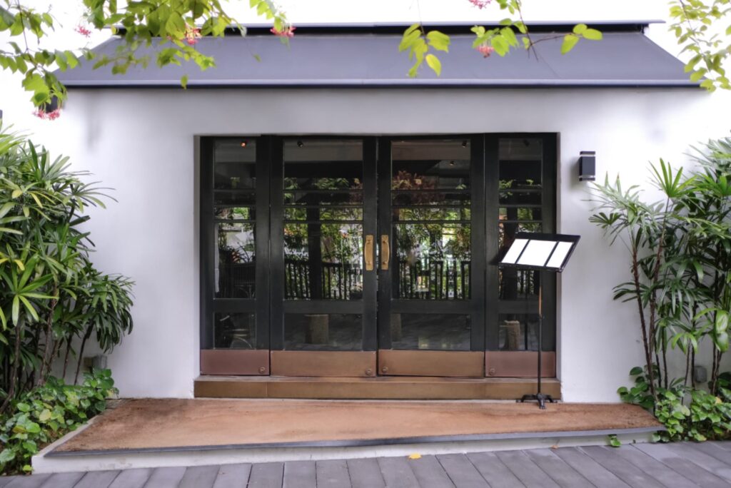 Restaurant doors surrounded by green plants, with a sample menu on a podium. The menu and doors are shaded by a large Calnova commercial awning