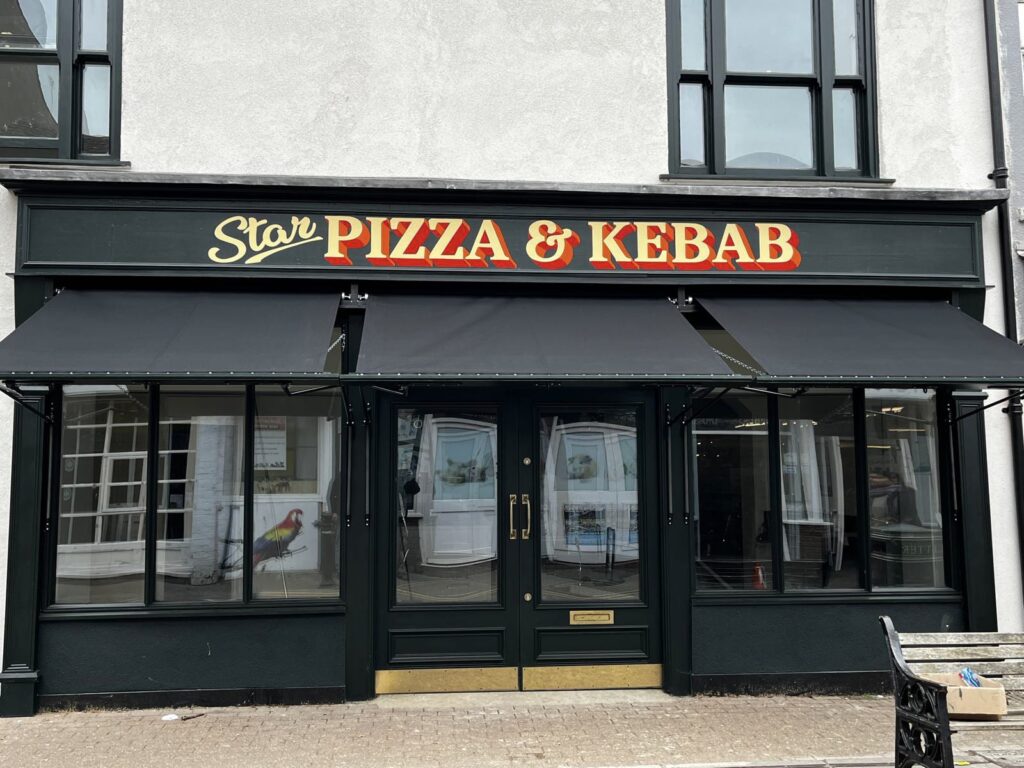 A shopfront painted with black accents and branding across the top that reads "Star Pizza & Kebab". Three black Victorian awnings are installed on the shopfront over the windows and beneath the signage.