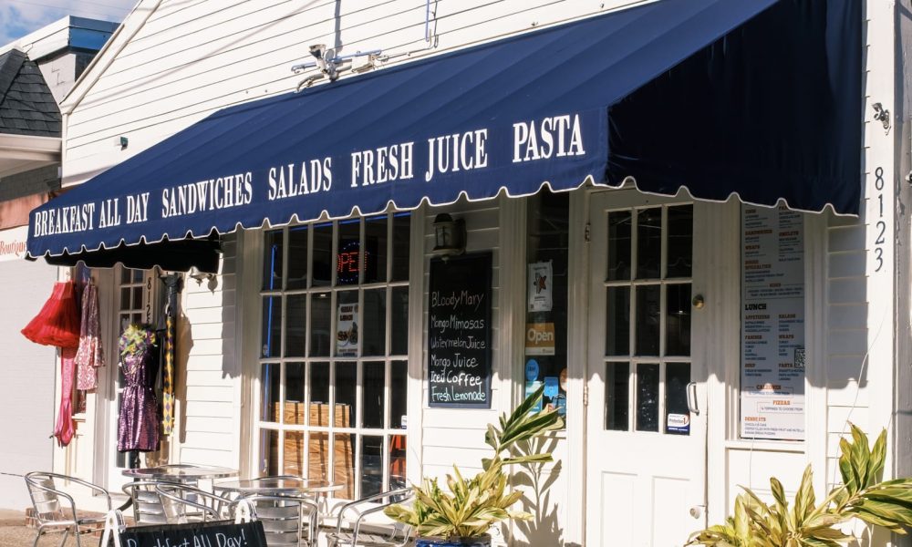 A white building with a large dark blue commercial canopy installed over the windows and doors. The commercial canopy has printed text on it describing the kind of food that business sells.